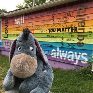 A stuffed Eeyore in front of a rainbow colored house with uplifting sayings and words